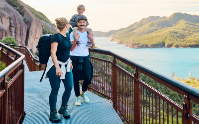 A family enjoying an outdoor hike on a scenic trail overlooking water and mountains. One adult carries the child on their shoulders while the other walks beside them.