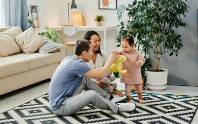 A family of 3 spending quality time together in a cozy living room. The parents are sitting on the floor, engaging with their young child who is holding a stuffed toy.