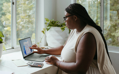 Woman working from home, using a laptop and calculator at a desk by the window, with plants in the background and natural light streaming in.