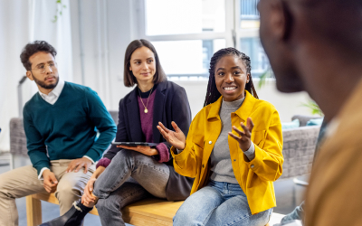 Diverse group of professionals sharing a laugh and chatting in a modern office setting, with one woman in a red blouse holding a tablet and smiling warmly.