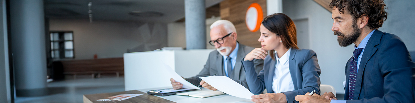 Three professionals in business attire seated at a table in a modern office, engaged in reviewing documents, with a staircase and architectural elements in the background.