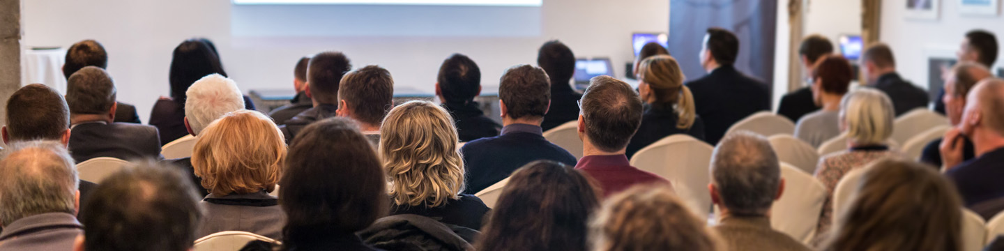 Audience seated in rows of white chairs in a conference room, attentively facing a presentation screen at the front