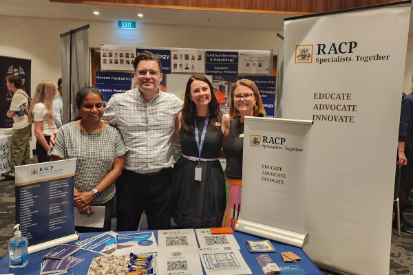 4 RACP representatives smiling at an information booth during a medical careers event. The table is covered with brochures, merchandise, and promotional material, with RACP banners displaying the words 'Educate, Advocate, Innovate' in the background.